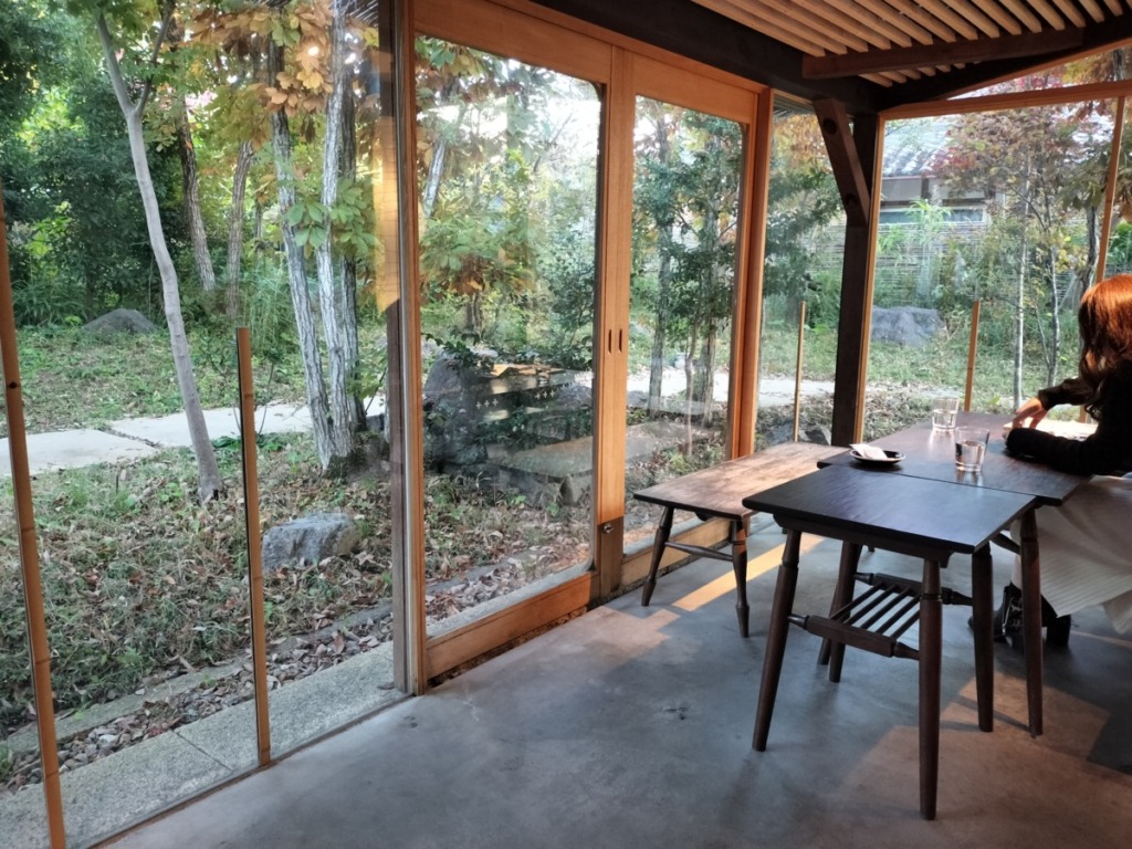 中島大祥堂 丹波本店 店内の様子。大きなガラス窓から庭の緑がよく見える、開放的で明るい空間。木製のテーブルと椅子が配置され、一人の客が窓の外を向いて座っている。