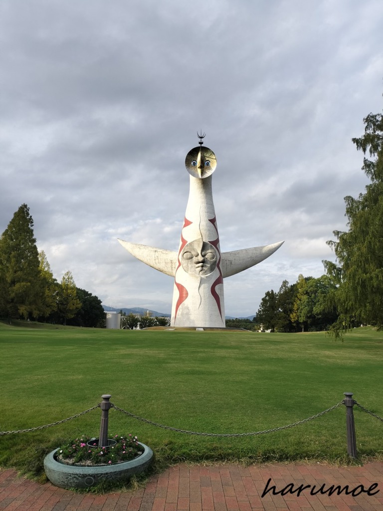 太陽の塔　広い芝生の中央に、岡本太郎が制作した白い巨大な塔『太陽の塔』が立っている。空は雲が広がり、塔の正面には赤い模様と中央の顔が見える。