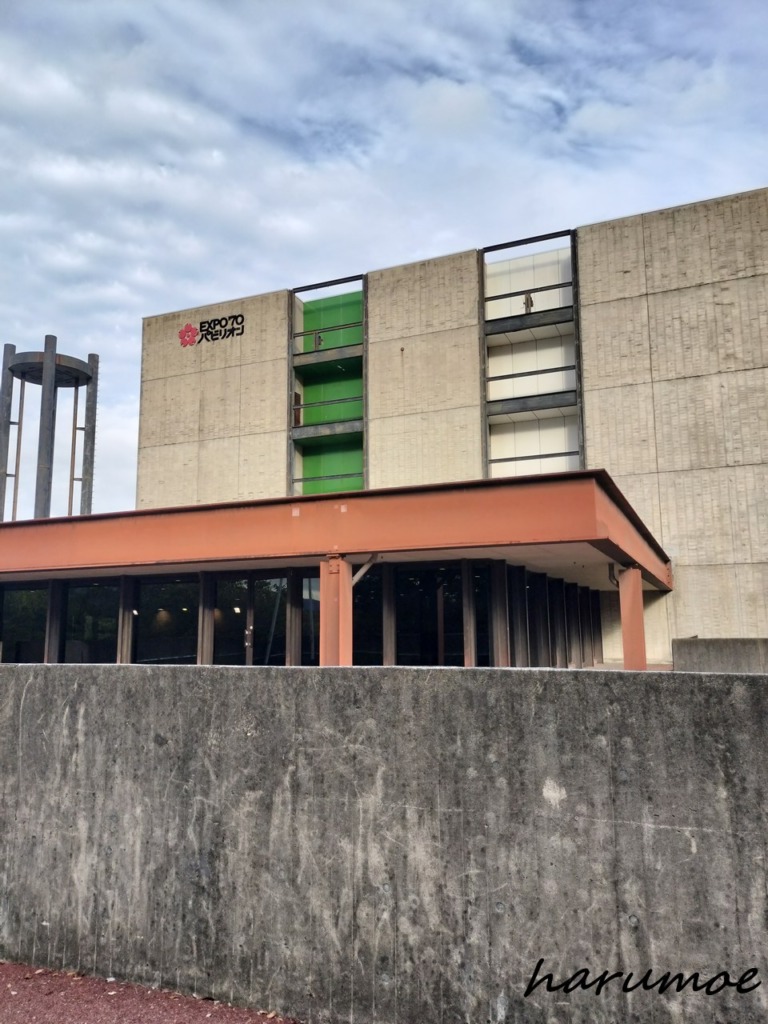 1970年大阪万博の資料館『EXPO’70パビリオン』の外観。コンクリートの建物に緑色の縦ラインが入り、上部にロゴが表示されている。前面には低いコンクリート壁がある。