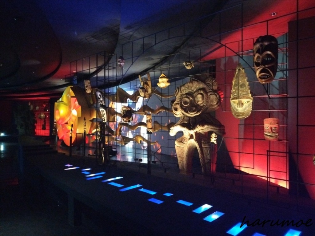 太陽の塔内部に広がる、薄暗い展示空間。中央には巨大な「生命の樹」の一部が見え、壁面には世界各地の仮面や彫刻のようなオブジェが、赤と青の照明で照らされながら展示されている。