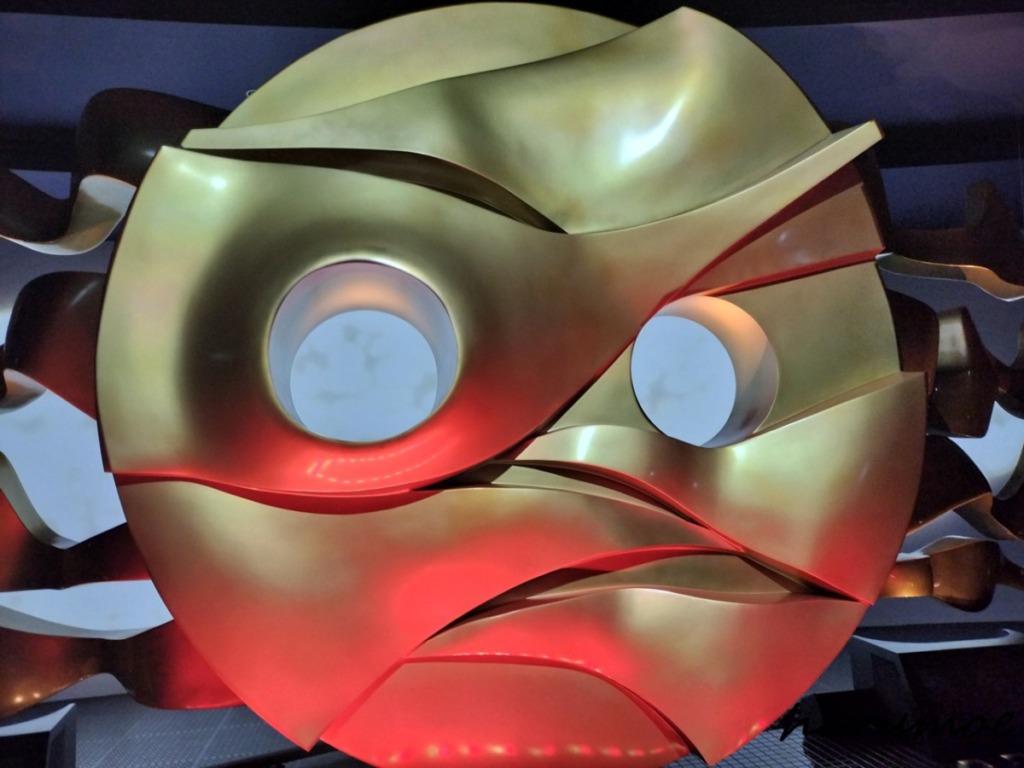 太陽の塔の内部にある、黄金色の抽象的な巨大な顔のオブジェ。湾曲したパーツが組み合わされ、目にあたる部分がくり抜かれている。下部は赤色の光で照らされている。