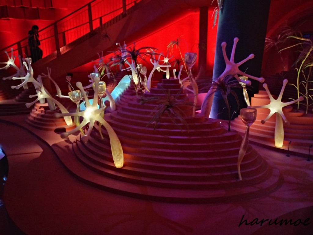 太陽の塔の内部、深紅の照明で照らされた空間。円形の階段状の展示台に、発光する触手状のオブジェや、植物のような奇妙な造形物が配置され、生命の根源を表現している。