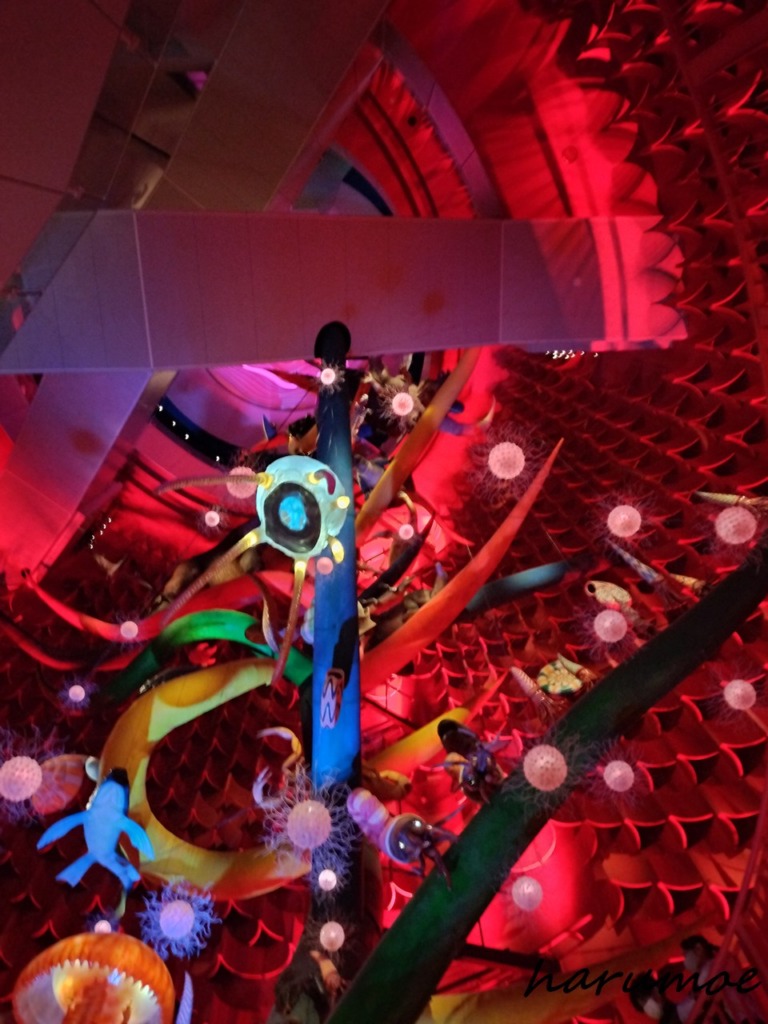 太陽の塔の内部を縦に貫く巨大な「生命の樹」の幹の部分を見上げた写真。幹には、古代魚、両生類、爬虫類などの生物模型が取り付けられ、周囲は深紅の照明と発光する球体の装飾で埋め尽くされている。