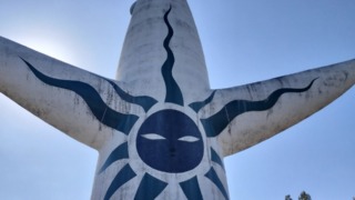 晴れた青空の下、太陽の塔を真下から見上げた写真。塔の背面にある、黒と青の模様で描かれた「黒い太陽」の顔が、全身に描かれている。