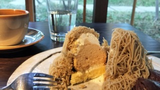 丹波栗のしぼりたてモンブランをナイフで切り分けた断面。モンブランペーストの内側には、生クリーム、メレンゲ、そして土台となるスポンジが見える。奥には水が入ったグラスとコーヒーカップ、窓の外の緑がある。