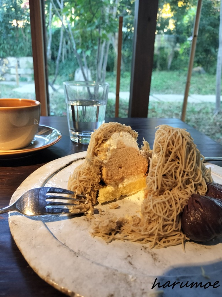 丹波栗のしぼりたてモンブランをナイフで切り分けた断面。モンブランペーストの内側には、生クリーム、メレンゲ、そして土台となるスポンジが見える。奥には水が入ったグラスとコーヒーカップ、窓の外の緑がある。